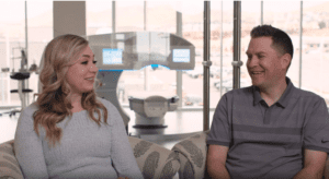 Rick and Chelsea Talbots, a white woman with blonde hair and white man with greying dark hair shown from the waist up sitting in arm chairs in front of the Waite Vision operating room.