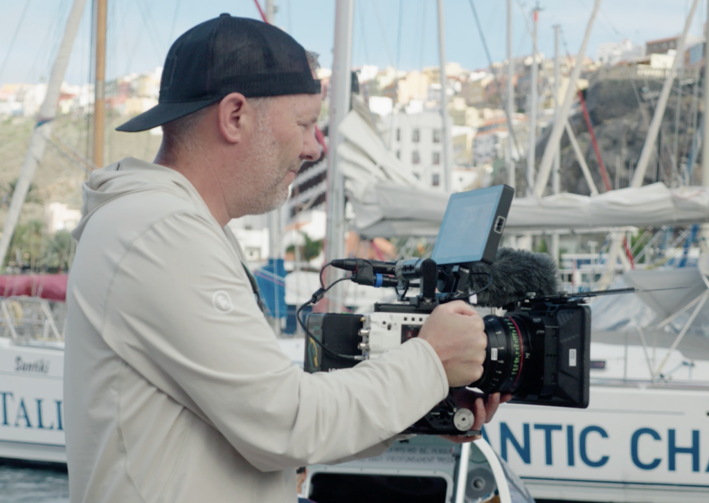 Tim Testimonial Photo; Tim is a white man in a white hoodie and a backwards ball cap shown from the waist up. He is holding a large camera pointed outward and looking intently at the screen. He is facing off to the right and standing in a boat marina