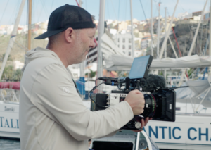 Tim Testimonial Photo; Tim is a white man in a white hoodie and a backwards ball cap shown from the waist up. He is holding a large camera pointed outward and looking intently at the screen. He is facing off to the right and standing in a boat marina