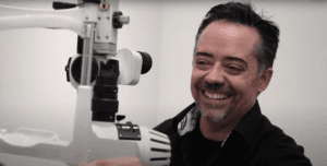 Dr. Podloski is shown from the shoulders up, sitting behind a vision test, ready to examine a patient's eye (out of view). He is a white man with dark, greying hair and greying scruff around his mouth. He is smiling.