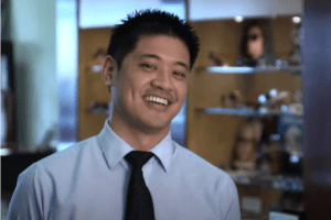 Dr Chiu testimonial - Dr Chiu is a man with light brown skin and spiky dark hair wearing a blue button up shirt and black tie. He faces the camera and has a big smile. Behind him are slightly blurry shelves covered in glasses displays