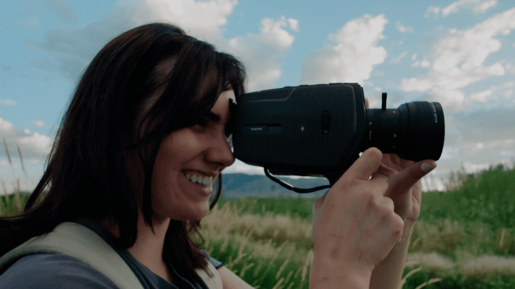 Jade, a young, brunette, white woman is shown from the shoulders up from her right side. She is wearing a blue-grey t-shirt and green backpack, and is holding a video camera up to her left eye. She is smiling, and her hands have small scratches on them.